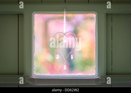 Un coeur ornement orné de cristaux se bloque sur une fenêtre avec un jardin coloré jeté dehors de la vue en arrière-plan. Banque D'Images