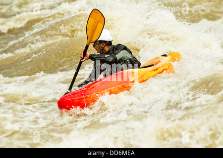 Un kayakiste dans un kayak rouge et orange manœuvre à travers des rapides d'eau vive mousseux et barattant. La personne porte un casque blanc, une combinaison sèche de protection noire, et tient une pagaie à lame jaune. Le Banque D'Images