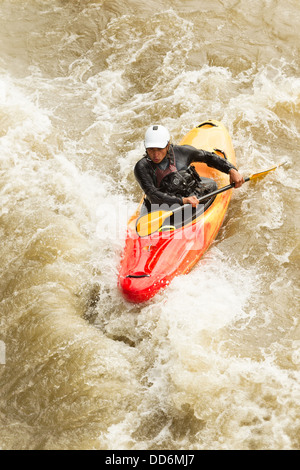 Un kayakiste dans un kayak rouge et jaune manœuvre à travers des rapides d'eau vive boueux et barattant. La personne porte un casque blanc, une combinaison de protection noire, et pagaie activement avec une lame jaune Banque D'Images