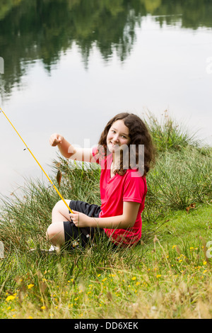 Photo verticale de jeune fille, à l'avant, la tenue de petits poissons qu'elle pris alors qu'à s'asseoir avec l'eau et arbres de retour Banque D'Images