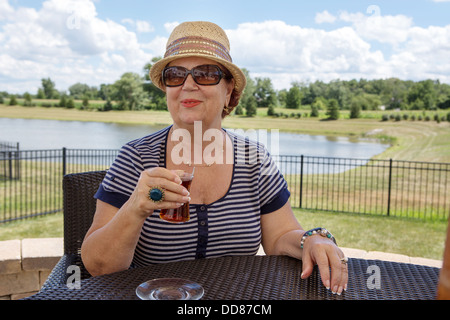 Senior femme élégante dans un chapeau et des lunettes de soleil en sirotant un verre dehors assis à une table avec vue sur un lac Banque D'Images