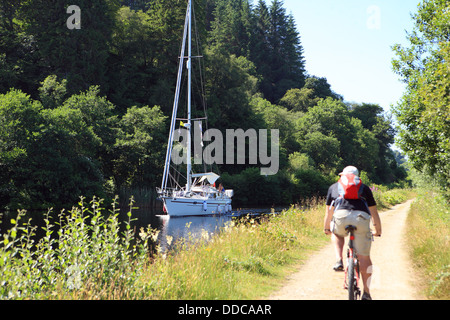 Cycliste sur la piste cyclable du Canal Crinan passant un yacht dans le canal à Argyll en Écosse Banque D'Images