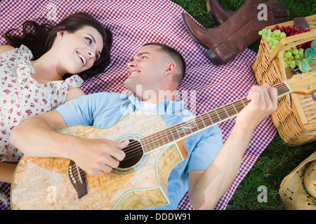 Happy Mixed Race Couple sur Couverture de pique-nique dans le parc et jouent de la guitare et chanter des chansons. Banque D'Images