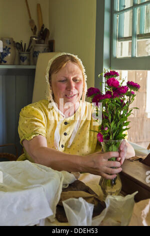 Old Bethpage, New York, États-Unis le 31 août 2013. JOANNE SMITH de Deer Park est de placer un vase de fleurs chrysanthème mauve sur la table de la cuisine à côté d'une fenêtre dans un bâtiment des années 1800 à Old Bethpage Village Restauration. La robe jaune et blanc dentelle hat elle porte sont American Civil War era clothing. Banque D'Images