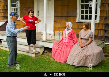 Old Bethpage, New York, États-Unis le 31 août 2013. PATRICIA JOSEPH, de College Point, en robe rouge, et jane HEILIG, de Melville, en robe taupe, parlez avec un homme et femme de visiter au cours de l'Olde Time Music Week-end à Old Bethpage Village Restauration, où la musique populaire des années 1800 et des danses de la guerre civile américaine est effectuée. Joseph & Heilig, vêtu d'American Civil War era style clothing et assis sur le porche de 12 Lawrence House, sont membres de l'ancien village de Bethphagé danseurs. Banque D'Images