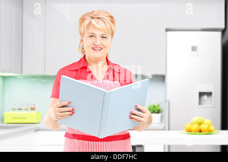 Smiling mature femme cuisinière tenant un livre de recettes dans la cuisine Banque D'Images