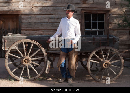 Cowboy debout devant un vieux wagon occidental et une grange portant un chapeau de cowboy noir, une chemise blanche, un Jean bleu et des chaps en cuir Banque D'Images