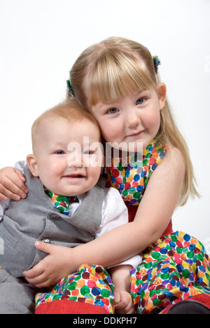 Petite fille et frère bébé assis ensemble à la fois câlins smartly dressed en costume et cravate et l'appariement spotty dress, UK Banque D'Images