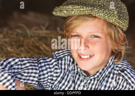 Young happy smiling blonde garçon enfant âgés de douze ou au début de l'adolescence portant chemise à carreaux & flat cap assis sur des bottes de paille ou de foin Banque D'Images