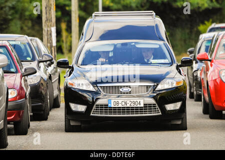 Un corbillard Ford noir se déplace lentement vers le bas une route étroite avec des voitures garées de chaque côté. Banque D'Images