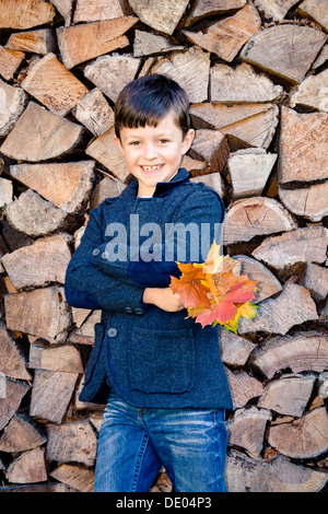 Boy holding Autumn Leaves, debout devant des bois empilés Banque D'Images