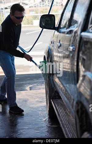 Un homme à l'aide d'un pinceau sur un tuyau pour nettoyer son véhicule à l'eau savonneuse dans un lave-auto. Banque D'Images