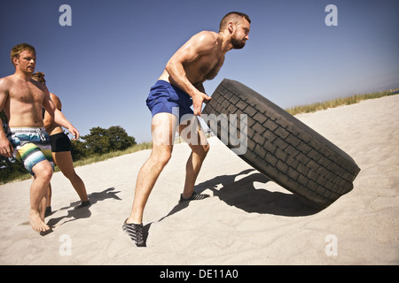 Athlète masculin difficiles renversant un pneu poids lourd. Des jeunes faisant l'exercice crossfit sur plage. Banque D'Images