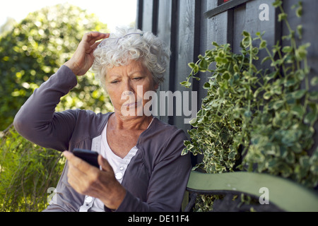 La femme assise sur un banc dans son jardin la lecture de message texte sur son téléphone portable Banque D'Images