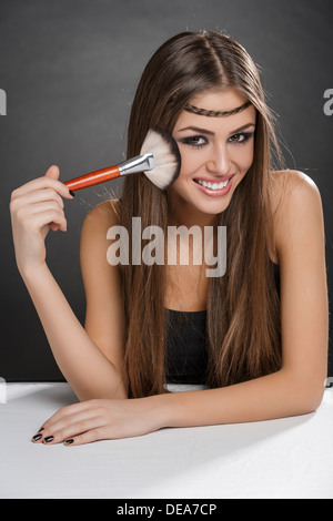 Portrait of a happy young woman applying makeup sur son visage avec un gros pinceau sur fond sombre. Banque D'Images