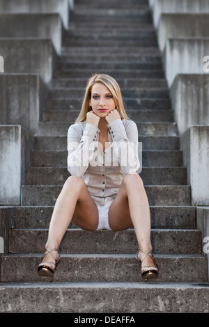 Jeune femme avec de longs cheveux blonds portant une chemise grise, un short blanc et de hauts talons assis sur des escaliers en pierre Banque D'Images