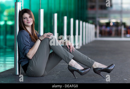 Jeune femme avec des talons hauts assis et posant devant un mur de verre vert Banque D'Images