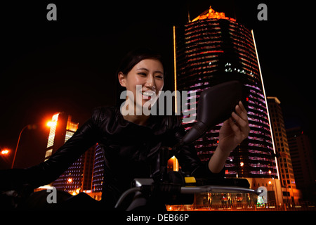 Belle jeune femme contrôle elle-même dans le miroir du côté d'une moto, à l'extérieur de nuit à Beijing Banque D'Images