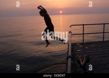 Soir d'été 2013 : A Young boy wearing wetsuit sautant hors d'une jetée dans la mer au coucher du soleil, Aberystwyth Wales UK Banque D'Images