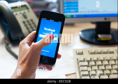 Woman's hand holding a smart phone avec facebook sur l'écran dans un bureau avec un bureau, téléphone, ordinateur écran et clavier Banque D'Images