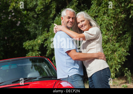 Smiling mature couple hugging against their red cabriolet Banque D'Images