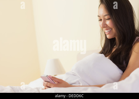 Happy young asian woman sitting in bed using smartphone Banque D'Images