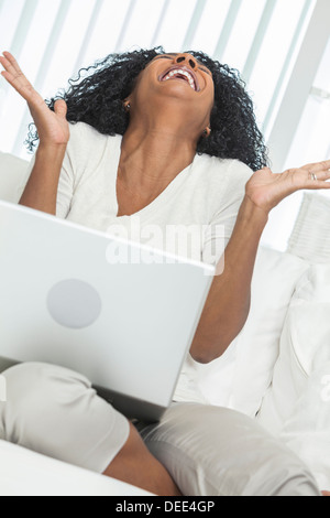 African American Woman à la maison assis sur un canapé ou fauteuil de célébrer et de rire en utilisant son ordinateur portable. Banque D'Images