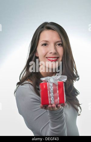 Jolie jeune femme aux longs cheveux brun et un beau sourire doux qui fait un cadeau rouge coloré pour un être cher Banque D'Images