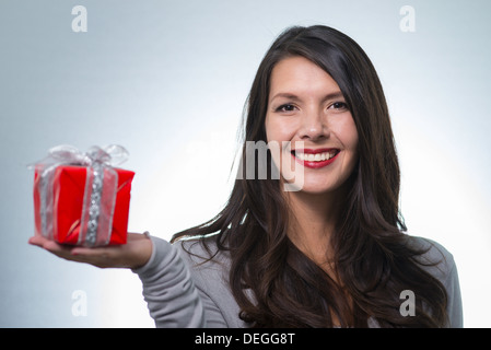 Jolie jeune femme aux longs cheveux brun et un beau sourire doux qui fait un cadeau rouge coloré pour un être cher Banque D'Images