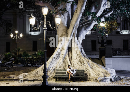 Homme assis sur un banc de la lecture sous un réverbère. Derrière lui, le tronc et les racines d'un ficus macrophylla, Banque D'Images