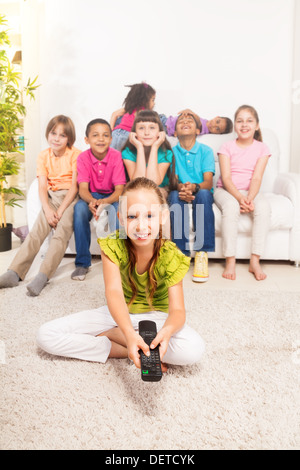 Petite fille avec la télécommande du téléviseur assis sur le plancher dans la salle de séjour avec ses amis assis dans le bus on background Banque D'Images