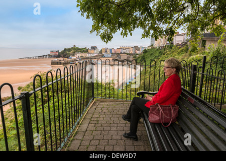 Femme plus âgée assise sur banc avec vue sur North Beach, Tenby Pembs.marée basse avec ses bateaux amarrés et terrasses des maisons de type Régence Banque D'Images