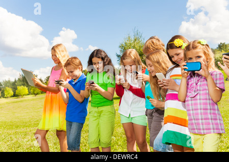 Grand groupe d'enfants occupés, les garçons et les filles à la recherche à leurs téléphones sms sms et jouant à l'extérieur Banque D'Images