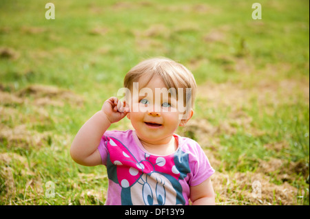 Baby sitting on grass holding her ear and smiling Banque D'Images