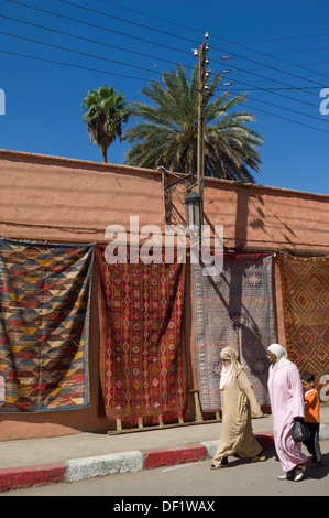 Les femmes en costume traditionnel marocain passé marche tapis suspendus à vendre, près de le palais de la Bahia, Marrakech, Maroc Banque D'Images