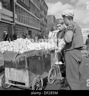 Photo historique, années 1950 par J Allan Cash, montrant un vendeur de rue afghan par sa petite charrette en bois à Kaboul, en Afghanistan, pesant des fruits pour un client. Un temps relativement pacifique pour le pays, cette époque a vu de nouveaux bâtiments construits, quelques pas faits vers la modernisation et faire du pays une société plus ouverte et prospère. Cela s'est terminé dans les années 1970 par une succession d'invasions, d'coups d'Etat et de guerres civiles. Banque D'Images