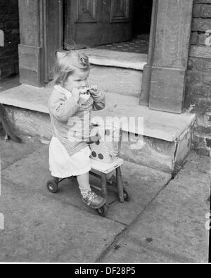 Photo historique de1950s d'une petite fille sur sa propre assise sur son cheval jouet en dehors d'une entrée de manger une glace lolly Banque D'Images