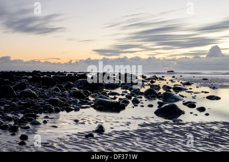 Une paisible plage de galets à marée basse avec des piscines de l'eau, big black de pierre mouillée et formes intéressantes dans le sable. Banque D'Images