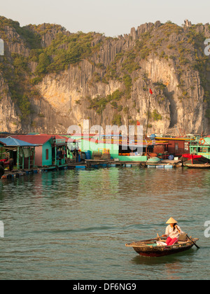 Une Vietnamienne dans un chapeau conique de lignes d'un bateau passé un village flottant au large de l'île de Cat Ba dans la baie de Lan Ha, Halong Bay, Vietnam. Banque D'Images