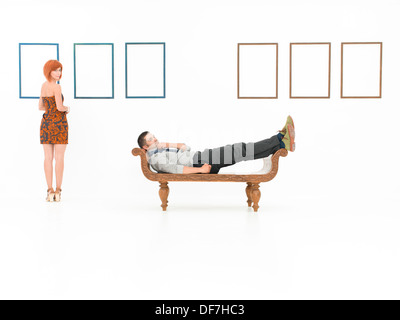 Young caucasian homme assis sur un banc en bois avec des gestes de pensée, dans une salle blanche, avec une femme et des cadres vides en arrière-plan Banque D'Images