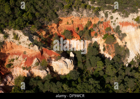 VUE AÉRIENNE.Falaise avec strates d'ocre multicolores en contraste saisissant avec la canopée environnante.Rustrel, Lubéron, Vaucluse, Provence, France. Banque D'Images