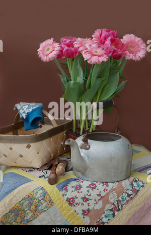 Un panier, un vieux pot de thé, gants, outils de jardinage, des tulipes roses, rose gerber daisies sur une courtepointe faite à la main. Banque D'Images