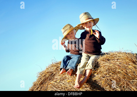 Deux enfants, un jeune garçon et son petit frère sont assis dehors sur des balles de foin, portant des chapeaux de paille sur une journée ensoleillée d'automne. Banque D'Images