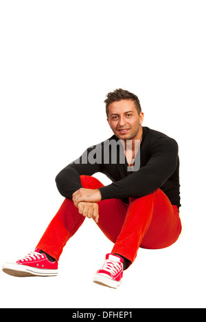 Modèle homme souriant en chemise noire et pantalon rouge assis sur le plancher contre fond blanc Banque D'Images