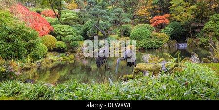Bronze Sculpture japonais grues paire par l'eau étang d'arbres plantes et arbustes à l'automne Panorama Banque D'Images