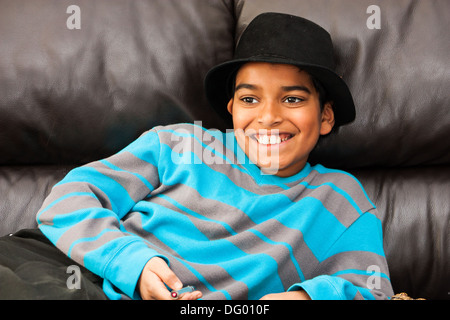 Un jeune garçon avec un grand sourire un sportif black fedora, allongé sur un canapé en cuir noir Banque D'Images