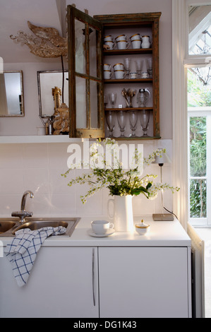 Verres sur des étagères au petit armoire en bois au-dessus de l'évier et plan de travail à pot de fleur blanche en blanc cuisine contemporaine Banque D'Images