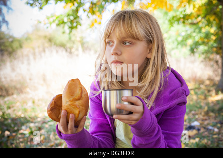 Petite blonde girl in autumn park mange de petites tourtes avec plateau Banque D'Images