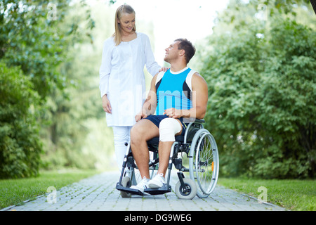 Jolie infirmière marcher avec male patient in a wheelchair in park Banque D'Images