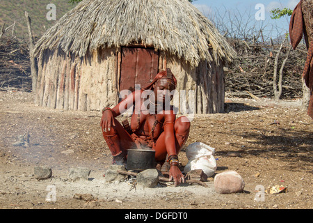 Jeune femme Himba, assis à une fosse de cuisson en face d'une hutte, Kaokoland, Kunene, Namibie Banque D'Images